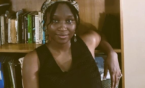 Amina Waya sitting in front of her bookshelf at home.